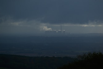A dark, overcast landscape features a distant industrial facility emitting smoke, situated beneath heavy clouds. The foreground consists of shadowy silhouettes of trees or bushes, while the horizon is barely visible due to the atmospheric haze.