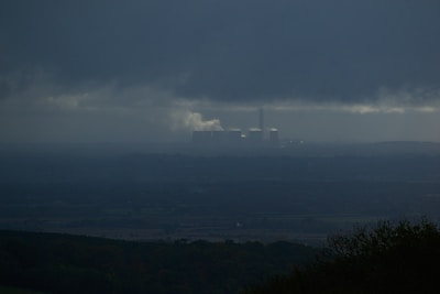A dark, overcast landscape features a distant industrial facility emitting smoke, situated beneath heavy clouds. The foreground consists of shadowy silhouettes of trees or bushes, while the horizon is barely visible due to the atmospheric haze.