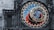 An ornate astronomical clock with intricate details and vibrant colors, featuring numbers, zodiac signs, and celestial designs. The clock is set against a stone wall, with statues and sculpted figures surrounding it.
