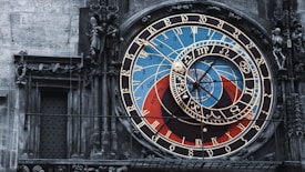 An ornate astronomical clock with intricate details and vibrant colors, featuring numbers, zodiac signs, and celestial designs. The clock is set against a stone wall, with statues and sculpted figures surrounding it.