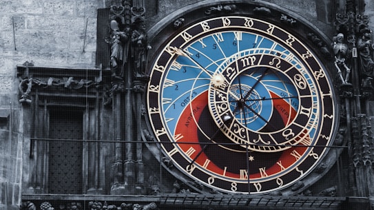 An ornate astronomical clock with intricate details and vibrant colors, featuring numbers, zodiac signs, and celestial designs. The clock is set against a stone wall, with statues and sculpted figures surrounding it.