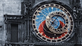 An ornate astronomical clock with intricate details and vibrant colors, featuring numbers, zodiac signs, and celestial designs. The clock is set against a stone wall, with statues and sculpted figures surrounding it.