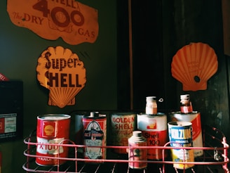 A collection of vintage oil cans is displayed on a shelf, featuring various old-fashioned Shell branding. The cans, which include Shell X-100 and Golden Shell Motor Oil, have a retro design with vibrant red and yellow colors. A backdrop of faded metal signs highlights the Shell brand with large logos and text.