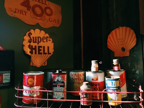 A collection of vintage oil cans is displayed on a shelf, featuring various old-fashioned Shell branding. The cans, which include Shell X-100 and Golden Shell Motor Oil, have a retro design with vibrant red and yellow colors. A backdrop of faded metal signs highlights the Shell brand with large logos and text.