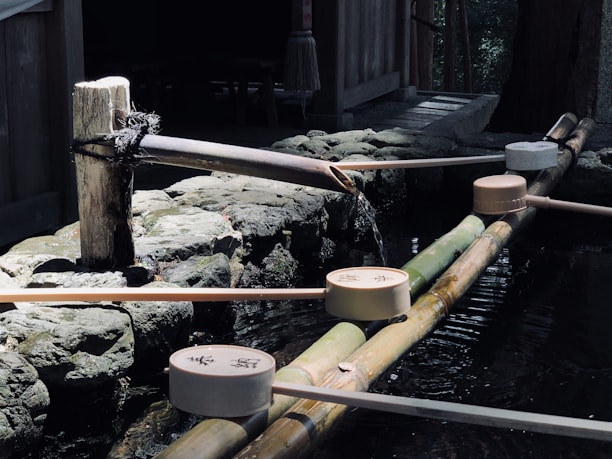 A traditional Japanese water purification fountain with wooden ladles positioned on a bamboo frame. Water is flowing gently from a spout, creating a tranquil atmosphere. Stones and wooden structures surround the area, indicating a serene outdoor setting.