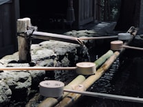 A traditional Japanese water purification fountain with wooden ladles positioned on a bamboo frame. Water is flowing gently from a spout, creating a tranquil atmosphere. Stones and wooden structures surround the area, indicating a serene outdoor setting.