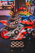 A row of arcade motorcycle racing games is lined up, featuring brightly colored machines with detailed graphics and logos. A person in the background stands near decorated countertops and a wall-mounted screen. The environment has a lively and playful ambiance, with vibrant colors and patterns.