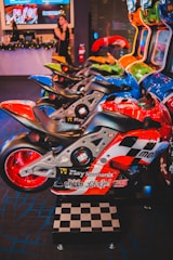 A row of arcade motorcycle racing games is lined up, featuring brightly colored machines with detailed graphics and logos. A person in the background stands near decorated countertops and a wall-mounted screen. The environment has a lively and playful ambiance, with vibrant colors and patterns.