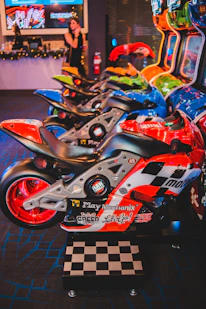 A row of arcade motorcycle racing games is lined up, featuring brightly colored machines with detailed graphics and logos. A person in the background stands near decorated countertops and a wall-mounted screen. The environment has a lively and playful ambiance, with vibrant colors and patterns.