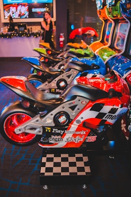 A row of arcade motorcycle racing games is lined up, featuring brightly colored machines with detailed graphics and logos. A person in the background stands near decorated countertops and a wall-mounted screen. The environment has a lively and playful ambiance, with vibrant colors and patterns.