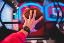 A hand wears a digital watch and holds a basketball, aiming towards a basketball hoop with vibrant neon lights in the background, creating a dynamic and energetic atmosphere.
