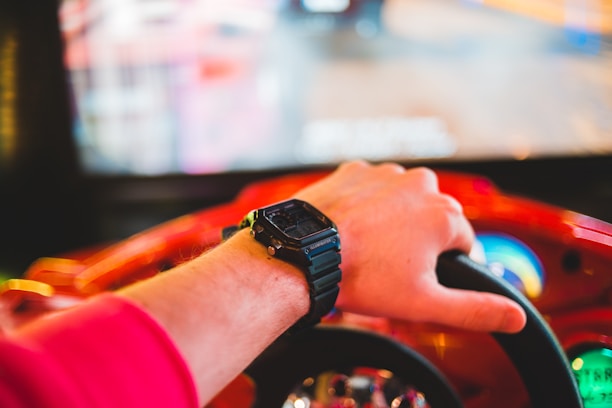 A person's hand wearing a black smartwatch is gripping the steering wheel of a red arcade racing game machine. In the background, there is a blurred screen displaying the game environment.