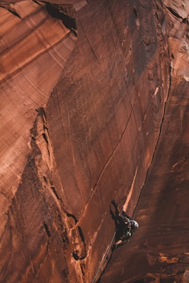 A climber is scaling a steep, rugged rock face. The rock surface features shades of orange and brown with distinct texture and layers. The climber is wearing a helmet and climbing gear, appearing focused on the ascent. The scene conveys a sense of challenge and adventure.