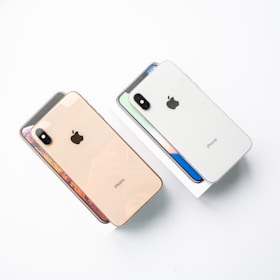 Two iPhones are placed side by side, each on top of its respective box. One phone has a gold back, and the other has a white back. Both feature a prominent logo and camera detailing, with the boxes showing images of the front screens.