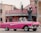 A classic pink convertible car is parked on a street in front of a pastel pink building with a sign reading 'La Floridita.' The building features vintage architectural details and flags on top. A person wearing a hat is seated inside the car. A tall, ornate lamppost stands nearby.