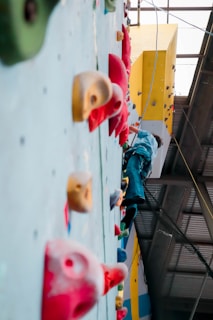 A person is climbing an indoor rock climbing wall with various colored holds. The scene is focused on the wall, showing different climbing holds in colors like red, yellow, and green. The climber is wearing dark pants and is attached to a rope for safety.