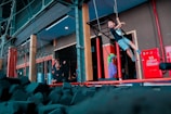 A young boy is swinging from ropes in an indoor adventure or play area. There are numerous foam blocks on the ground, likely for safety. Nearby, adults and possibly another child are observing the activity near a red hydrant box.