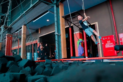 A young boy is swinging from ropes in an indoor adventure or play area. There are numerous foam blocks on the ground, likely for safety. Nearby, adults and possibly another child are observing the activity near a red hydrant box.
