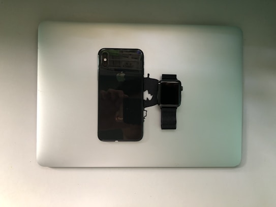 A smartphone and a smartwatch are placed on top of a closed laptop, all of which are identifiable as Apple products.