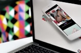A laptop has a vibrant abstract background displayed behind it, consisting of a colorful geometric pattern. A smartphone is positioned in front of the laptop, showing an image with tropical elements and a play button, possibly indicating a video or media content.