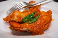 A close-up view of a dish featuring a piece of lobster covered in a red sauce, garnished with green leaves. A silver fork and knife are placed adjacent to the lobster on a white plate.