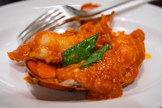 A close-up view of a dish featuring a piece of lobster covered in a red sauce, garnished with green leaves. A silver fork and knife are placed adjacent to the lobster on a white plate.