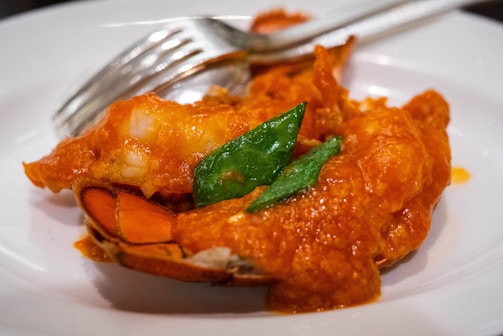 A close-up view of a dish featuring a piece of lobster covered in a red sauce, garnished with green leaves. A silver fork and knife are placed adjacent to the lobster on a white plate.