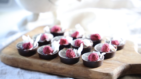 A wooden board featuring an arrangement of chocolate cups filled with cream and topped with bright red raspberries, accompanied by white decorative elements.