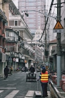 A city street scene with a person in a reflective vest cleaning the road. The street is lined with older, traditional-style buildings alongside modern skyscrapers in the background. Several bicycles are parked on the right and a cart is being pushed by someone in the center of the road. Overhead, numerous power lines and cameras can be seen, and the sky appears overcast.
