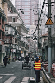 A city street scene with a person in a reflective vest cleaning the road. The street is lined with older, traditional-style buildings alongside modern skyscrapers in the background. Several bicycles are parked on the right and a cart is being pushed by someone in the center of the road. Overhead, numerous power lines and cameras can be seen, and the sky appears overcast.