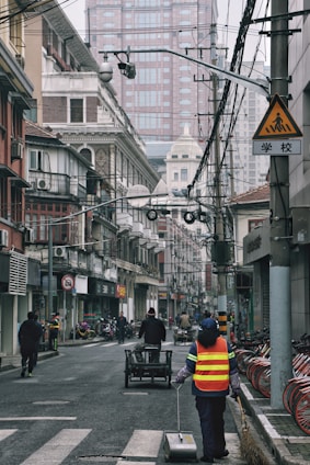 A city street scene with a person in a reflective vest cleaning the road. The street is lined with older, traditional-style buildings alongside modern skyscrapers in the background. Several bicycles are parked on the right and a cart is being pushed by someone in the center of the road. Overhead, numerous power lines and cameras can be seen, and the sky appears overcast.