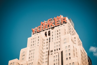 A tall Art Deco style skyscraper with the words 'NEW YORKER' prominently displayed in large red letters at the top against a clear blue sky.