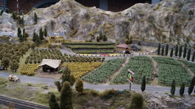 A miniature landscape features a rural setting with fields of crops arranged in neat rows. There is a small farmhouse with a red roof, a barn, and a few scattered buildings nestled among trees. The scene includes a winding road with a vintage car, hills in the background, and a billboard displaying vegetables.