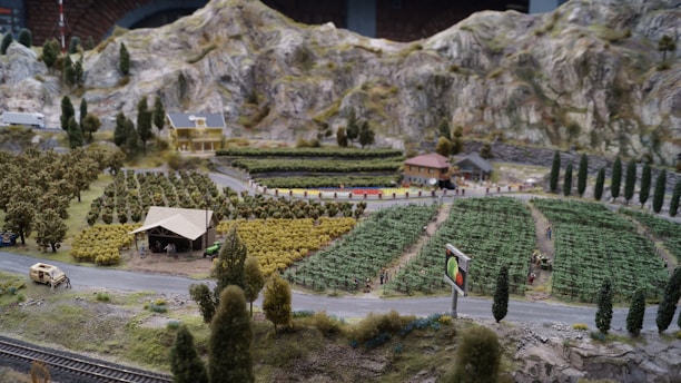 A miniature landscape features a rural setting with fields of crops arranged in neat rows. There is a small farmhouse with a red roof, a barn, and a few scattered buildings nestled among trees. The scene includes a winding road with a vintage car, hills in the background, and a billboard displaying vegetables.