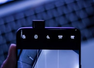 A smartphone with its front camera elevated above the screen displaying various icons including volume mute, aspect ratio 4:3, flash settings, HDR auto, and another mute icon. The background features an out-of-focus keyboard with blue lighting, giving a tech-centric ambiance.