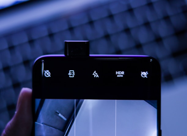 A smartphone with its front camera elevated above the screen displaying various icons including volume mute, aspect ratio 4:3, flash settings, HDR auto, and another mute icon. The background features an out-of-focus keyboard with blue lighting, giving a tech-centric ambiance.