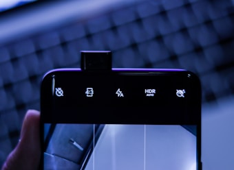 A smartphone with its front camera elevated above the screen displaying various icons including volume mute, aspect ratio 4:3, flash settings, HDR auto, and another mute icon. The background features an out-of-focus keyboard with blue lighting, giving a tech-centric ambiance.
