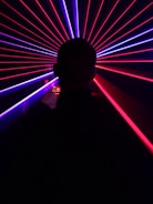 A silhouette of a person is in the foreground, facing an array of vibrant red and blue laser beams radiating outward, creating a dynamic and futuristic visual effect.