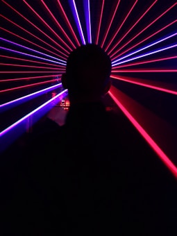 A silhouette of a person is in the foreground, facing an array of vibrant red and blue laser beams radiating outward, creating a dynamic and futuristic visual effect.