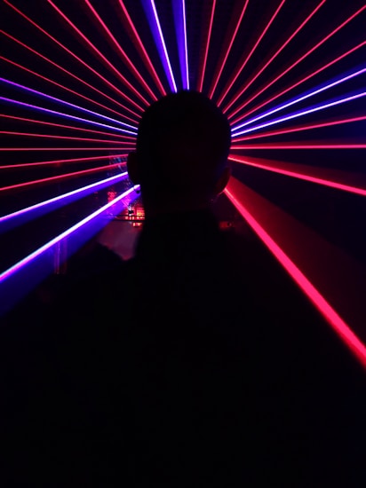 A silhouette of a person is in the foreground, facing an array of vibrant red and blue laser beams radiating outward, creating a dynamic and futuristic visual effect.