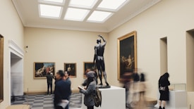 A museum gallery with a central sculpture of a human figure on a pedestal surrounded by framed paintings on the walls. People are walking and observing the art, with some taking photos and others reading or writing notes. The room has a calm and spacious atmosphere, with soft lighting from the ceiling.