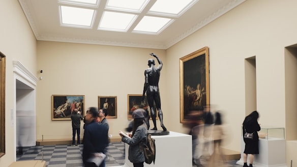 A museum gallery with a central sculpture of a human figure on a pedestal surrounded by framed paintings on the walls. People are walking and observing the art, with some taking photos and others reading or writing notes. The room has a calm and spacious atmosphere, with soft lighting from the ceiling.