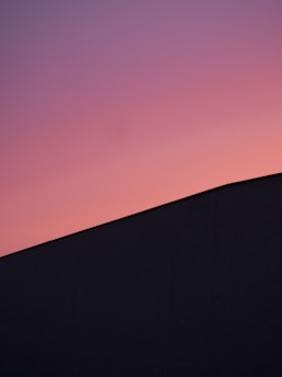 A minimalistic landscape with a gradient sky transitioning from pink to purple above a dark, silhouetted structure with a sloping roof.