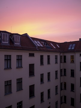 A multi-story residential building with a traditional red-tiled roof is bathed in the soft hues of a sunset. The building has numerous windows, some of which reflect the warm colors of the sky. A small silhouette of a bird is visible against the sky, enhancing the tranquil mood.