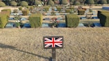 A cemetery with neatly arranged gravestones and well-maintained hedges. There are rose bushes with blooms scattered among the headstones. A small plaque in the foreground features the Union Jack, indicating a connection to the United Kingdom.