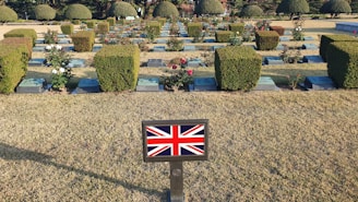 A cemetery with neatly arranged gravestones and well-maintained hedges. There are rose bushes with blooms scattered among the headstones. A small plaque in the foreground features the Union Jack, indicating a connection to the United Kingdom.
