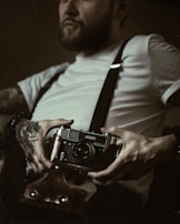 A person with tattoos on their arm holding a vintage camera, wearing a white shirt and suspenders. The image has a moody and artistic feel, with soft lighting and muted tones.