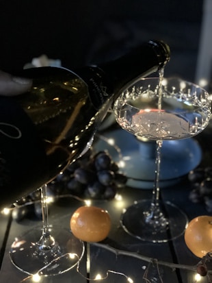 A bottle of sparkling wine is being poured into an elegant crystal glass. Surrounded by a dark setting, the scene includes grapes and oranges placed on the table, along with soft, glowing string lights that add a festive ambiance.