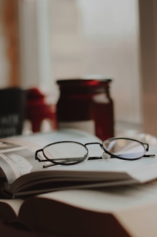 A pair of eyeglasses resting on an open book, with a blurred background that includes a dark red jar. Soft focus and warm lighting add to the cozy ambiance.