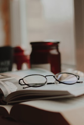 A pair of eyeglasses resting on an open book, with a blurred background that includes a dark red jar. Soft focus and warm lighting add to the cozy ambiance.